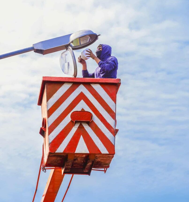 Asian electrician in bucket boom truck is repairing street lamppost against blue sky