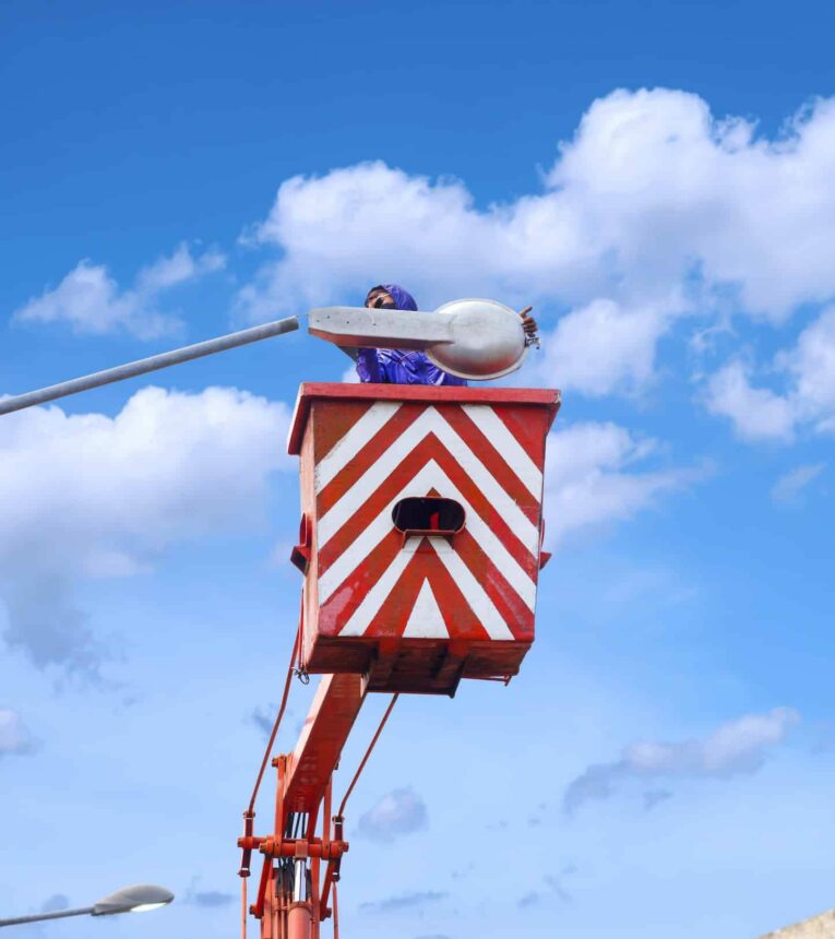 Electrician in bucket boom truck is repairing street light post against white clouds on blue sky