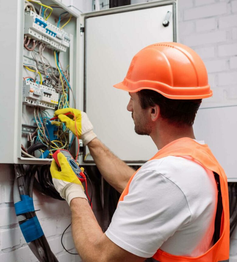 Handsome electrician in hardhat and gloves holding multimeter near electrical distribution box