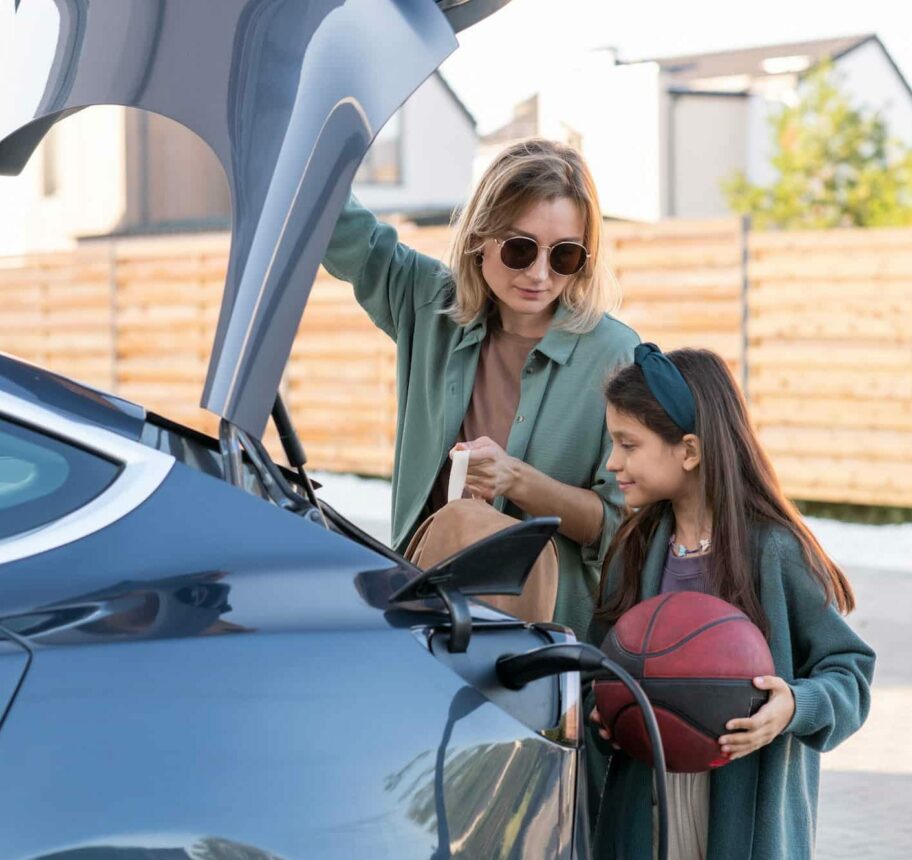 Little girl and young woman standing by trunk of electric car