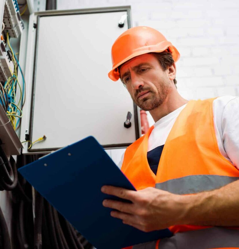 Low angle view of handsome electrician holding clipboard near electrical distribution box