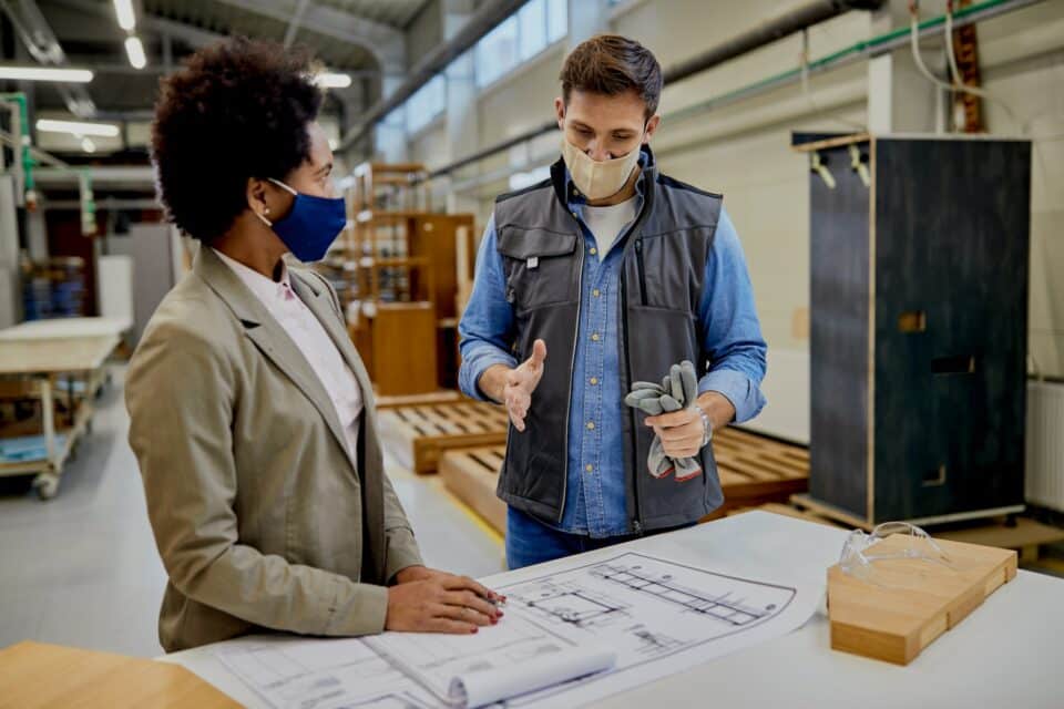Male carpenter explaining design plans to African American female manager at production facility.