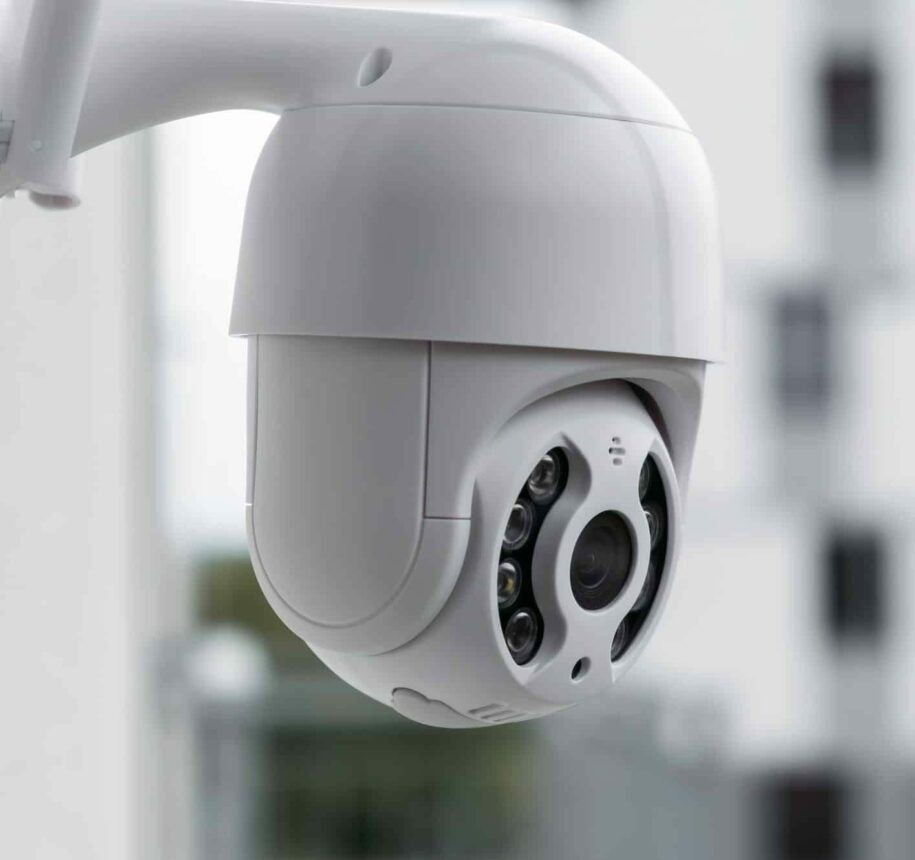 Modern CCTV camera on a wall with a blurred multi-storey building on the background.